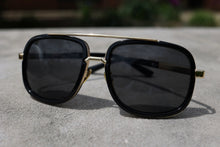 Load image into Gallery viewer, Black aviator-style sunglasses with gold metal frames and dark lenses, resting on a textured gray concrete surface with a blurred outdoor background.